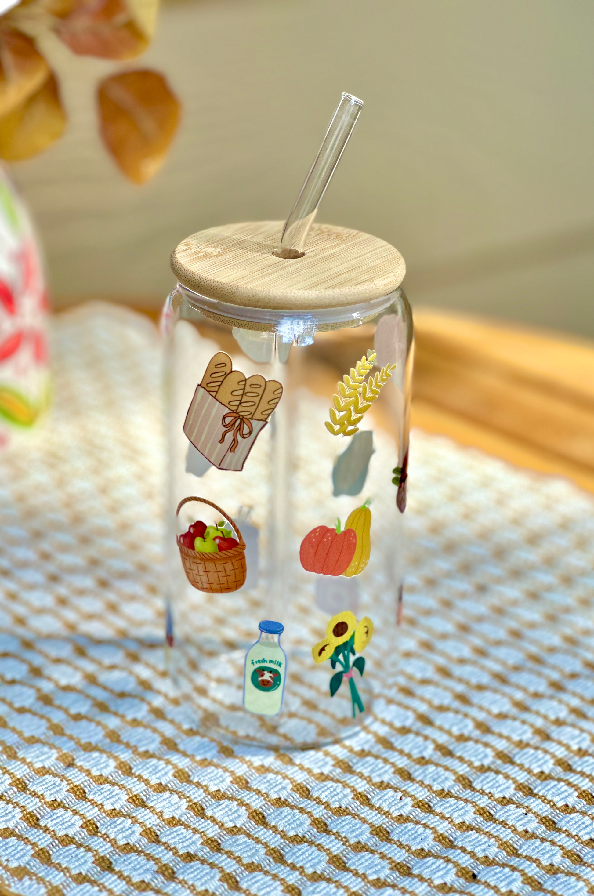 Clear tumbler with wooden lid and straw, featuring illustrations of everything you will find at a farmers market- pumpkins, flowers, bread, milk.